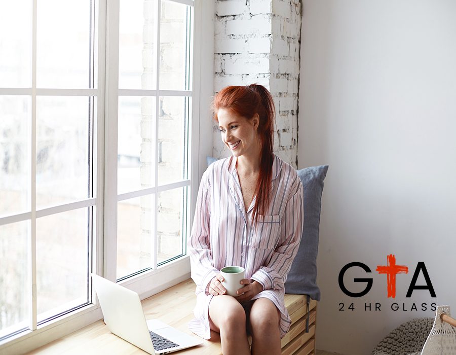 Beautiful young female with cute smile and ginger hair in ponytail enjoying slow morning at home, sitting by large window, drinking coffee, surfing internet on laptop. Daylight sunshine, soft colors