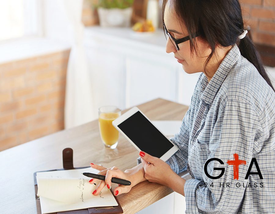 Cropped shot of serious businesswoman manages finances, holds tablet computer, looks attentively in notebook, sits at wooden kitchen desk, drinks orange fresh juice. Lifestyle, technology concept