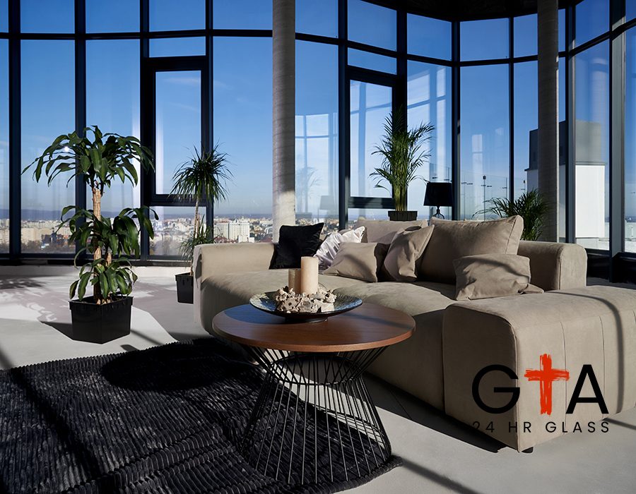 Front view of fashionable interior spacious room with large panoramic window with incredible view on city and cozy grey sofa with pillows and green flowerpots. Concept of modern interior.