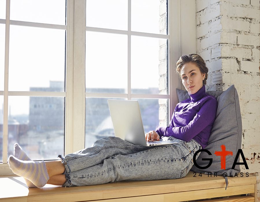 Young woman blogger in stylish clothes sitting on windowsill with pillow behind her back, having pensive thoughtful look while working on new article or post for her blog on social media, using laptop